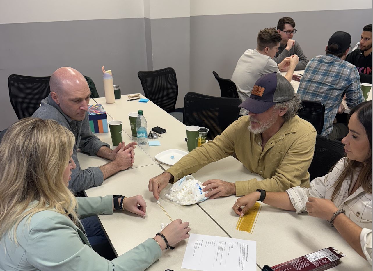 Coworkers leaning in around a table, building a spaghetti and marshmallow structure together during a team development workshop