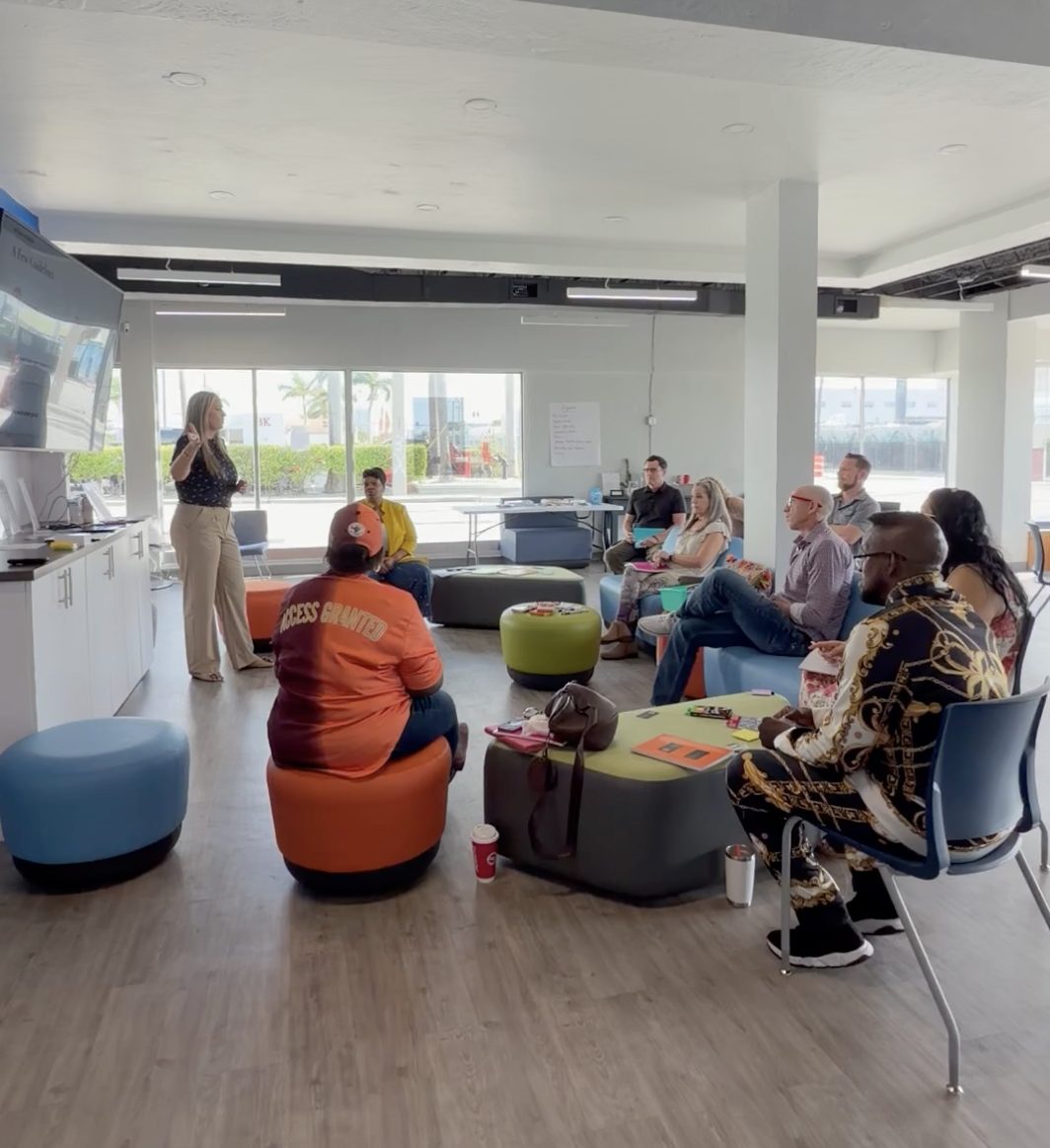 Nicole Meyer standing and facilitating a team workshop while participants seated on colorful ottomans and chairs listen