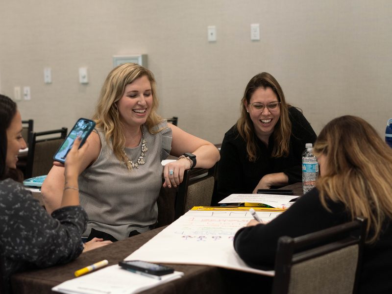 Four workshop participants laughing together at a table while one writes on a large sticky-note chart during a group activity